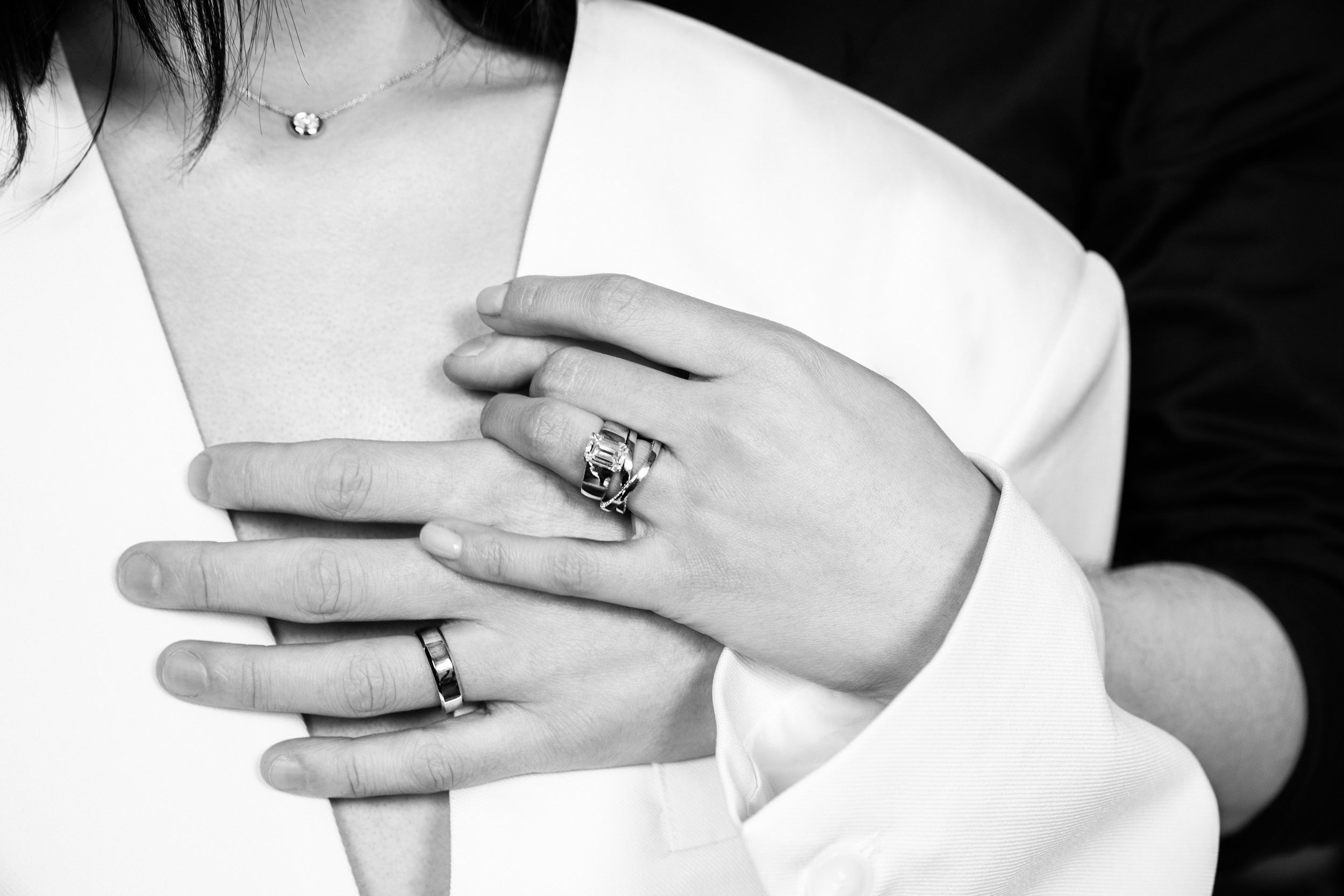 a model iwth dark hair in a white jacket is wearing a custom engagement diamond ring with an emerald diamond and a crossover solid 14k gold wedding band. Paired with her husbands hand wearing a simple custom gold band. Design a custom bridal or engagement ring with Sophie Ratner. Learn more or book an appointment.