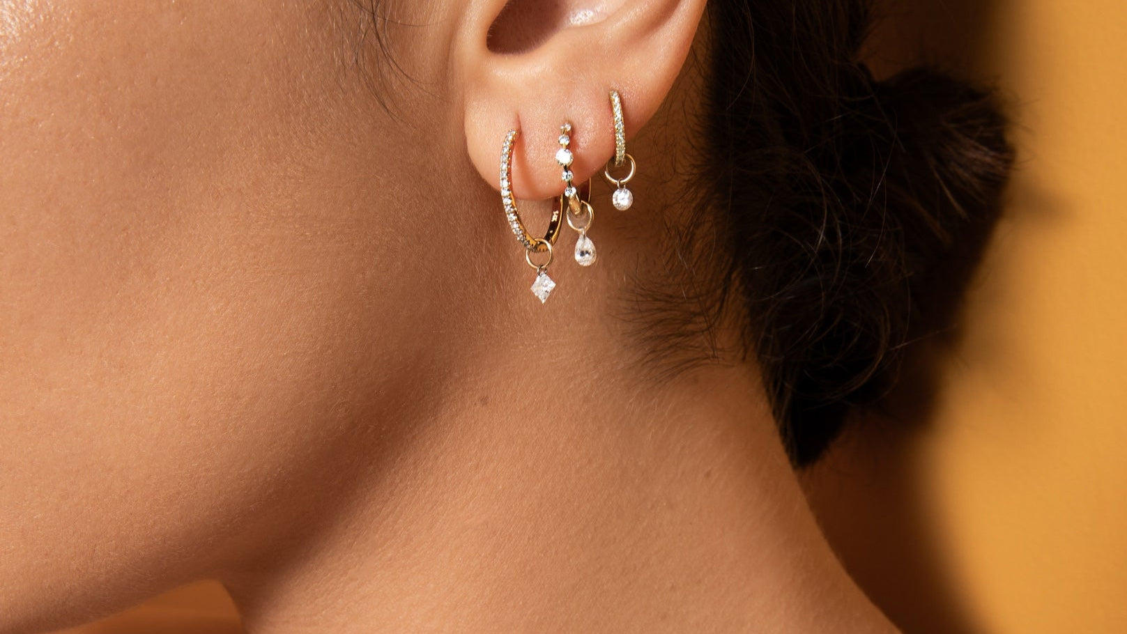 Close-up of a person wearing gold hoop earrings and a delicate gold necklace against a warm-toned background.