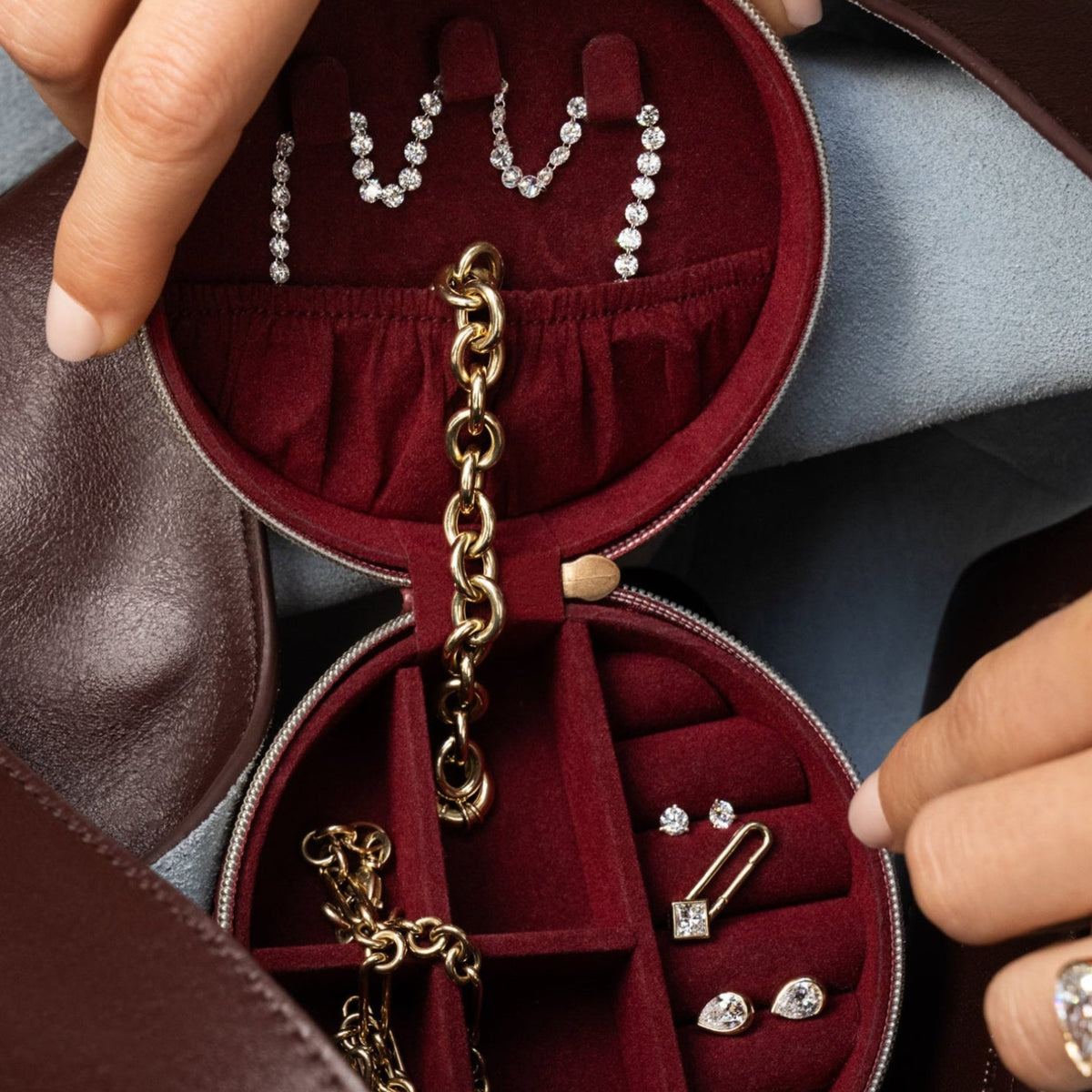 An open round burgundy jewelry organizer pouch inside a brown leather bag, holding various gold and silver jewelry pieces including earrings, necklaces, and rings. A person's hands with gold rings are holding the organizer.