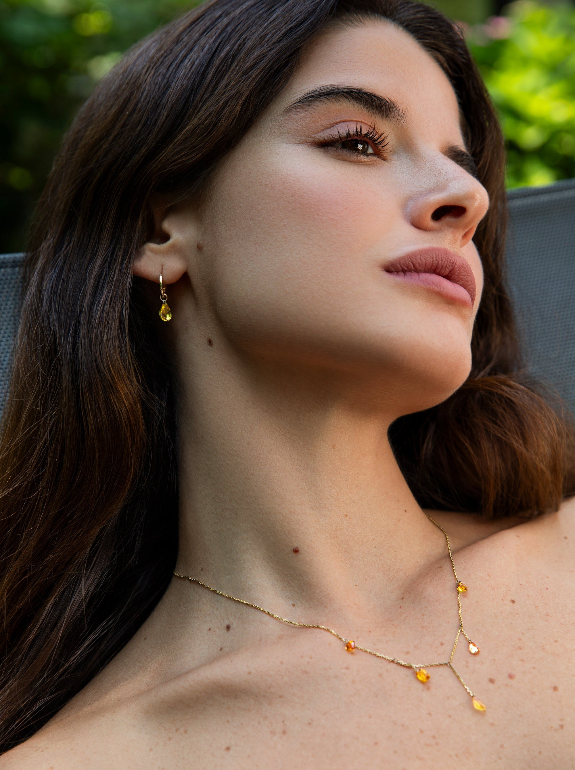 A close-up lifestyle shot of a woman wearing delicate gold jewelry, featuring a fine Y-shaped necklace with small gold charms and matching drop earrings.