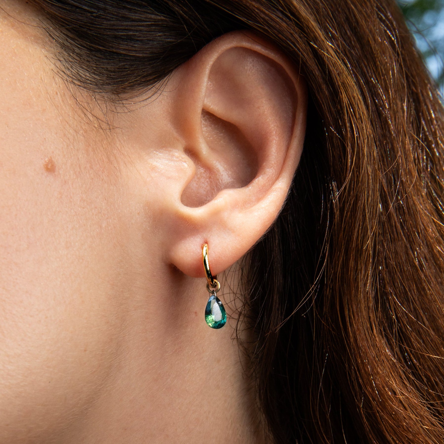A close-up of a person's ear wearing a small gold hoop earring with a pear-shaped green sapphire charm.