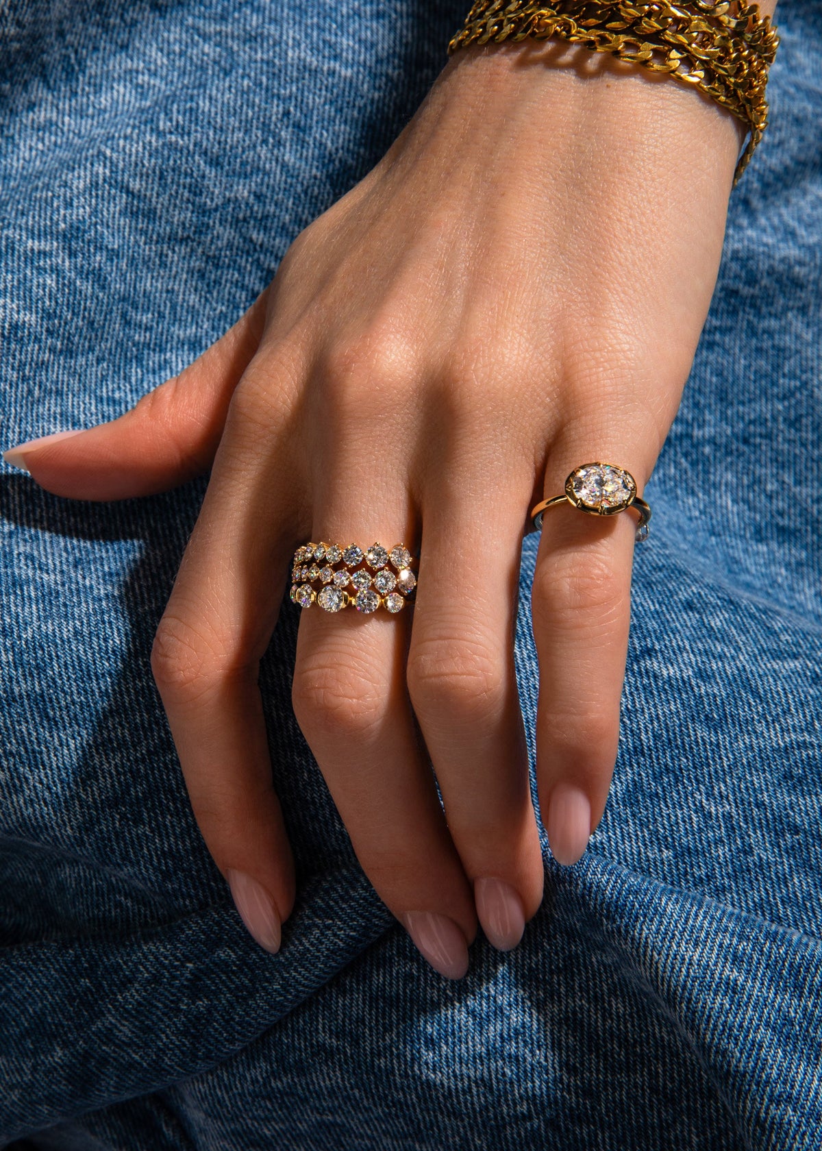 A close-up of a person's hand resting on blue denim, showcasing two gold rings with clear gemstones. One ring features three stacked bands of round-cut stones, and the other has a larger oval-cut stone.