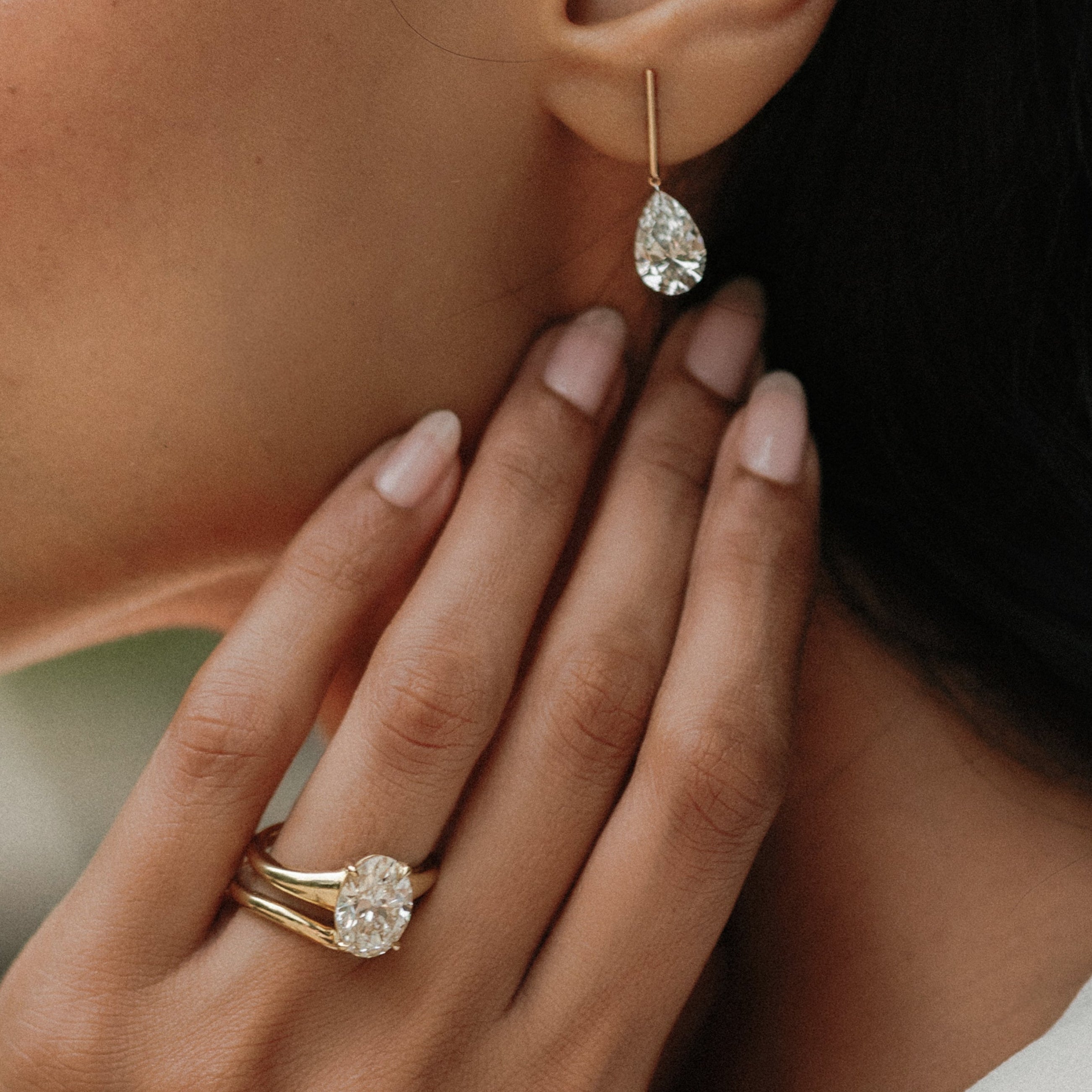 A close-up lifestyle shot of a woman wearing a delicate gold teardrop dangle earring and a matching gold band ring with a round-cut clear gemstone.