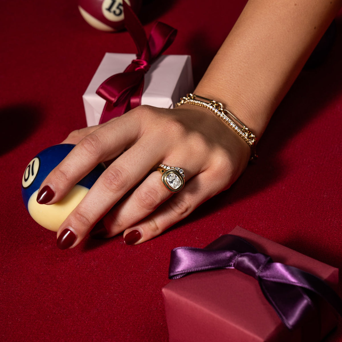 A model wearing the victorian engagement ring and wedding band holds a cue ball on a red felt pool table with gift boxes. Celebrating the sophie ratner gift guide, shop now. 