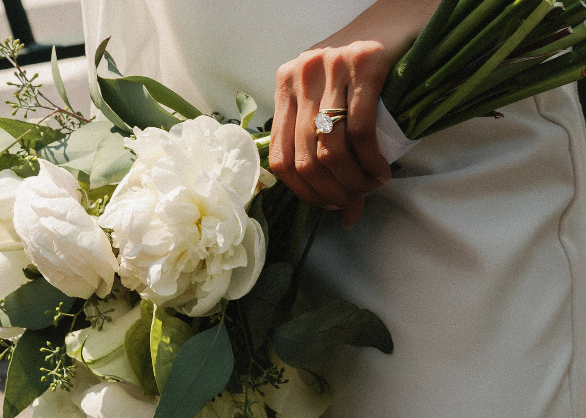 A close-up lifestyle shot of a bride in a white satin wedding dress holding a white peony bouquet with green stems, wearing a silver ring on her finger.
