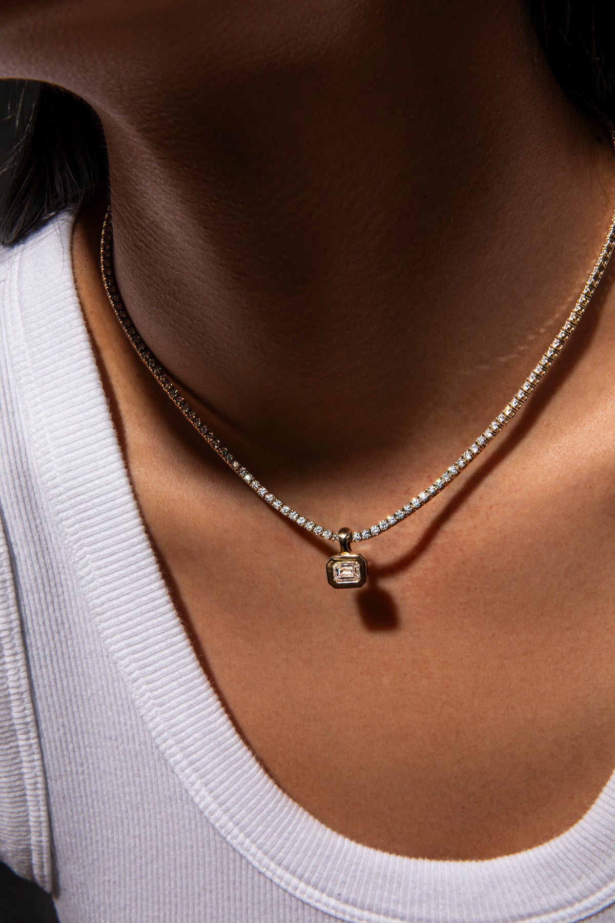 A close-up of a person's neck and upper chest wearing a delicate gold chain necklace with a small square pendant set with clear stones, paired with a white ribbed tank top.