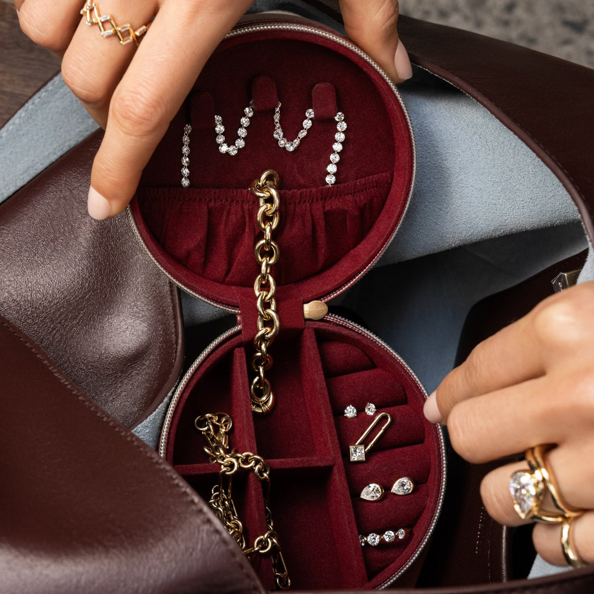 An open round burgundy jewelry organizer pouch inside a brown leather bag, holding various gold and silver jewelry pieces including earrings, necklaces, and rings. A person's hands with gold rings are holding the organizer.