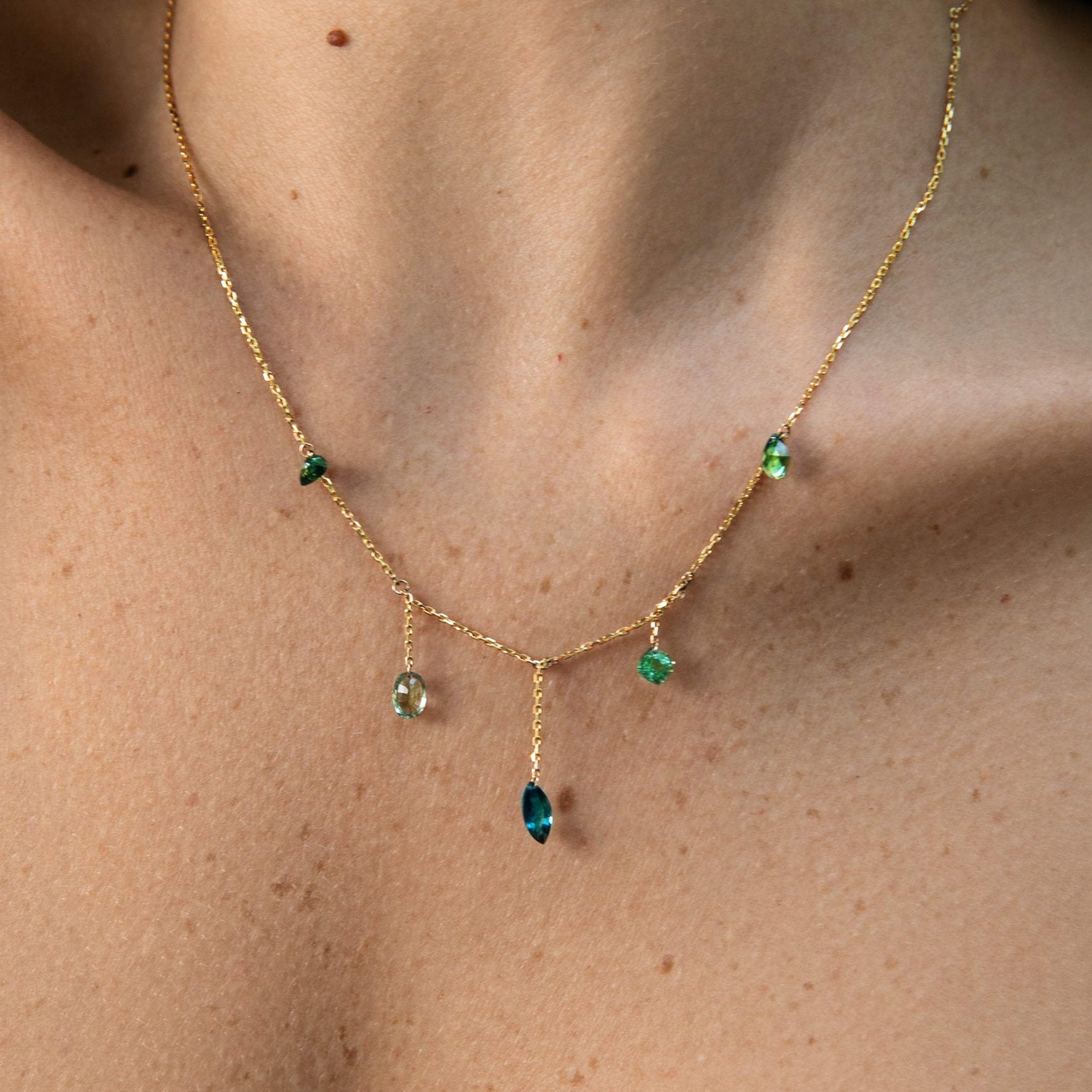 A delicate gold necklace with small green gemstone accents and a central teardrop-shaped blue-green stone, shown worn on a person's neck and upper chest.