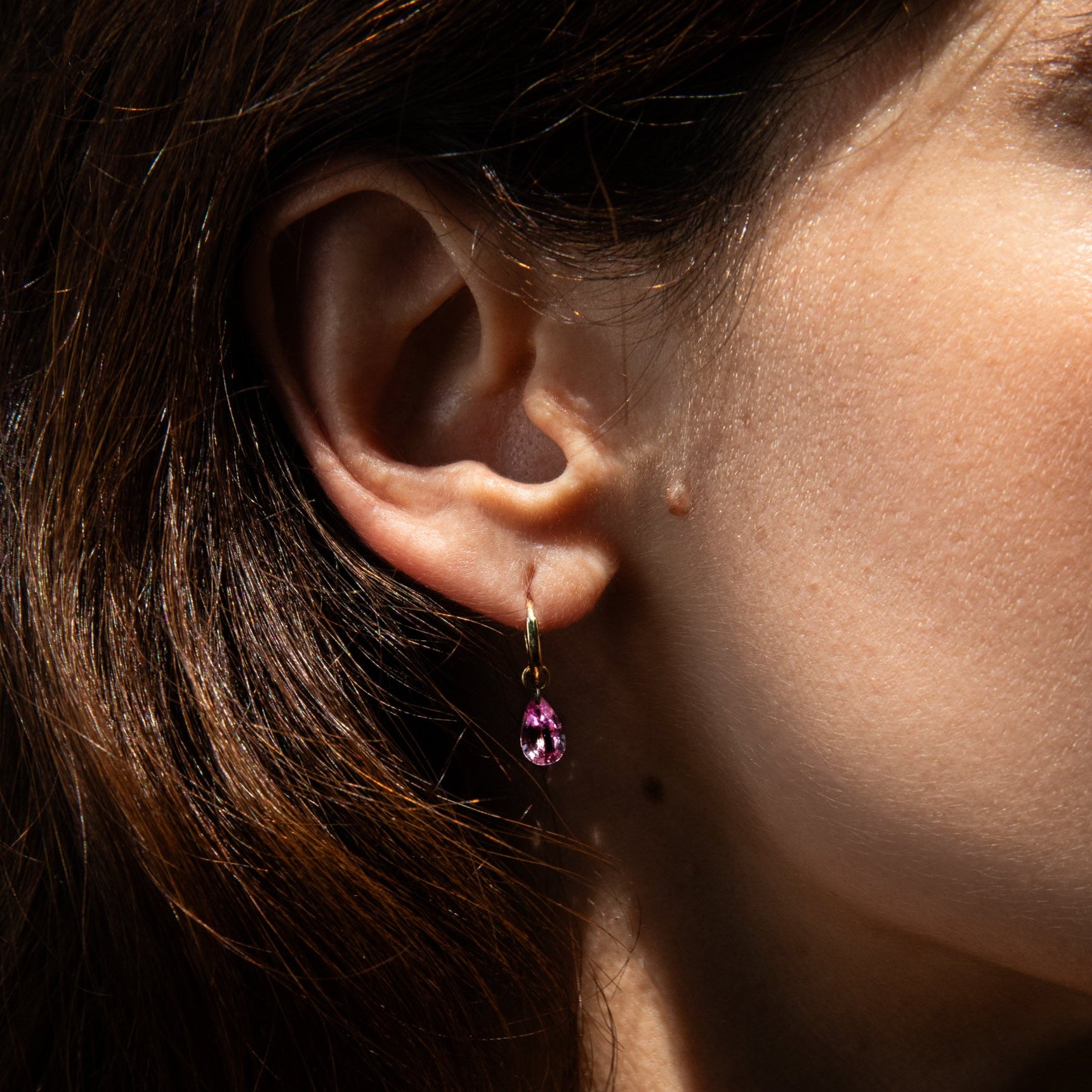 A close-up side profile of a woman wearing the 8mm gold huggie paired with a pear cut pink sapphire charm for huggies. set against her dark brown hair and warm skin under natural sunlight.