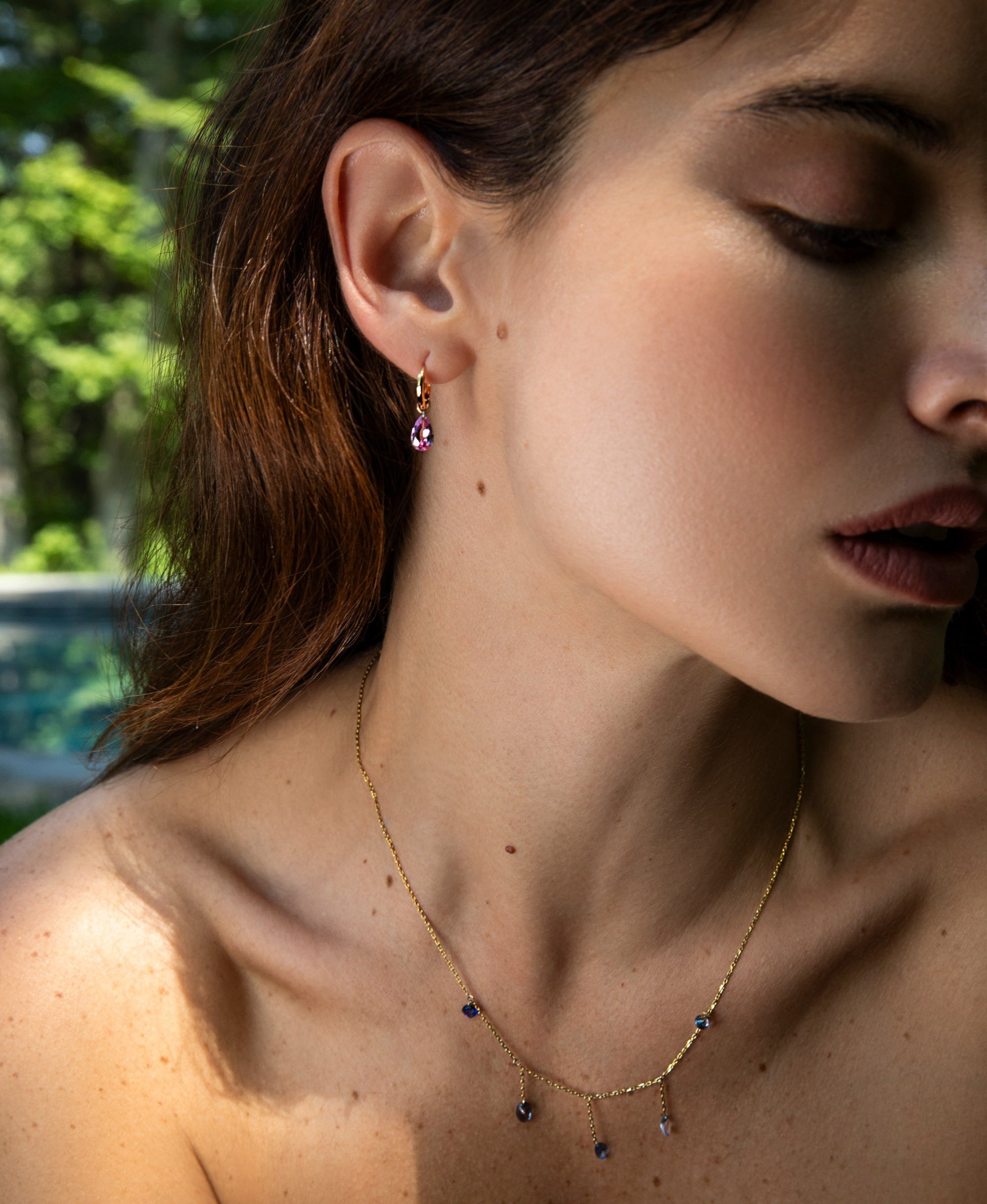 A close-up lifestyle shot of a woman wearing a delicate gold necklace with small dark blue and purple dangling charms, along with matching drop earrings, set against a blurred outdoor background.