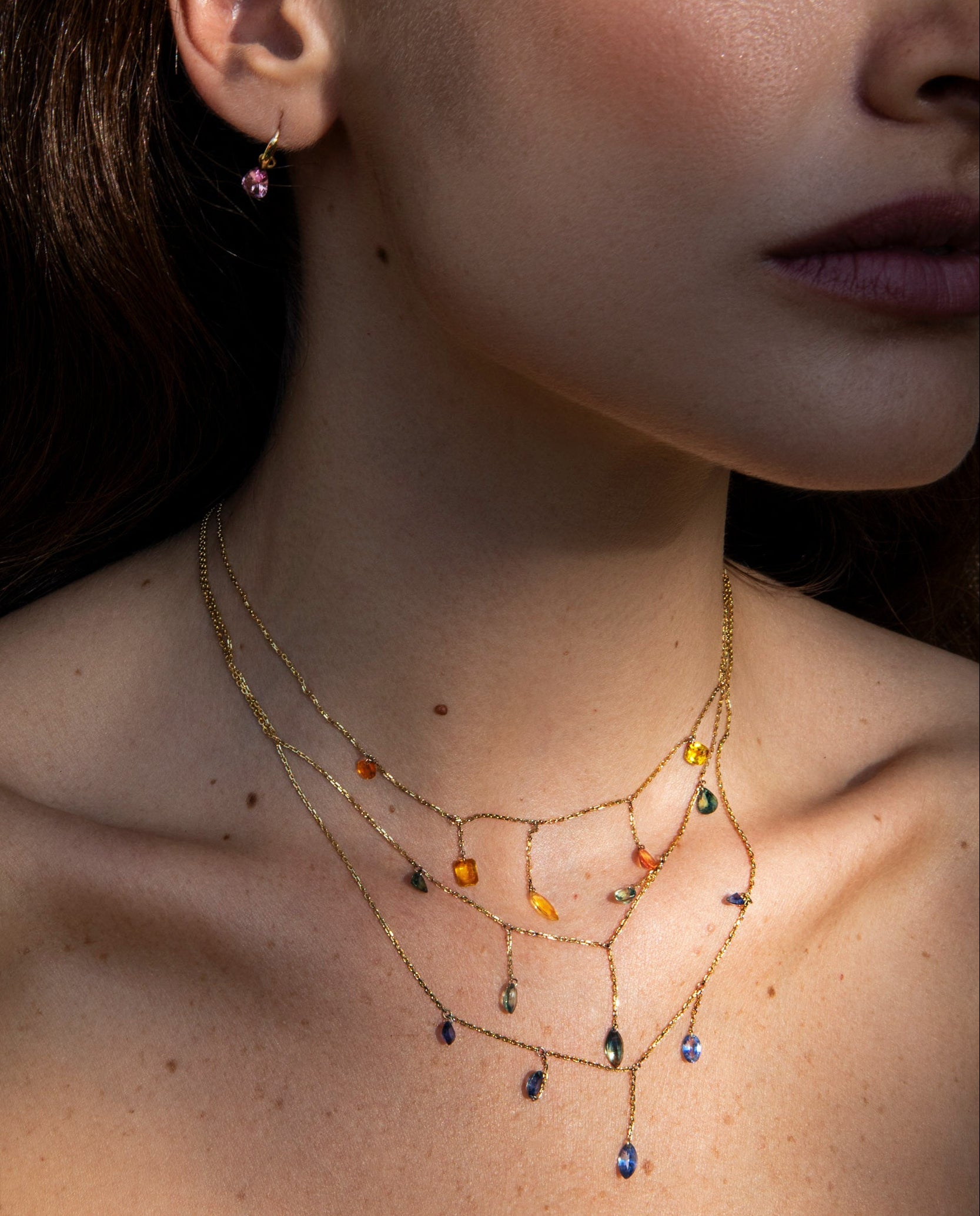 A close-up lifestyle shot of a model wearing a delicate gold layered necklace with small amber and blue gemstone accents, along with matching dangling earrings.