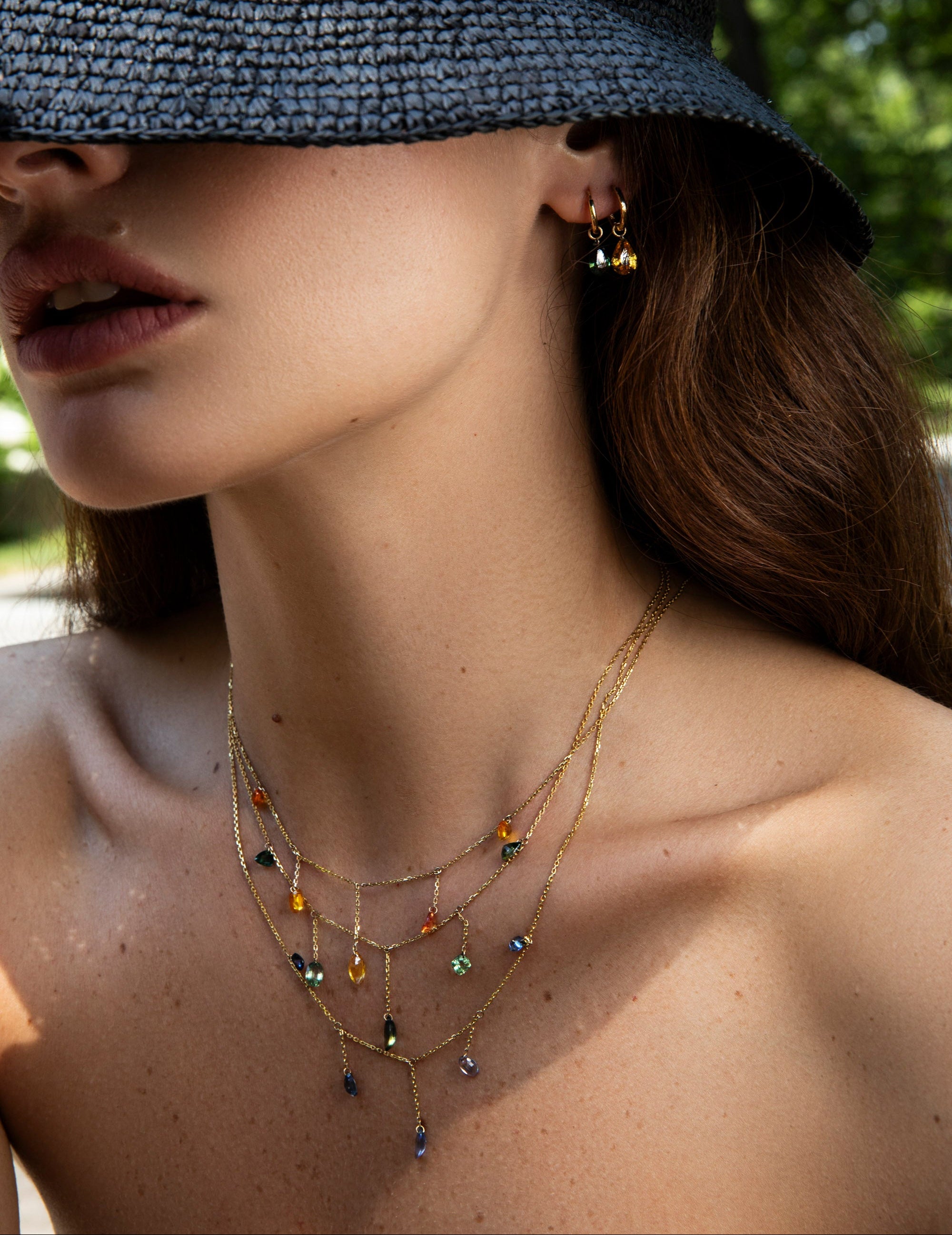 A close-up lifestyle shot of a model wearing a delicate gold multi-layered necklace with small colorful gemstone pendants, along with a black woven sun hat.