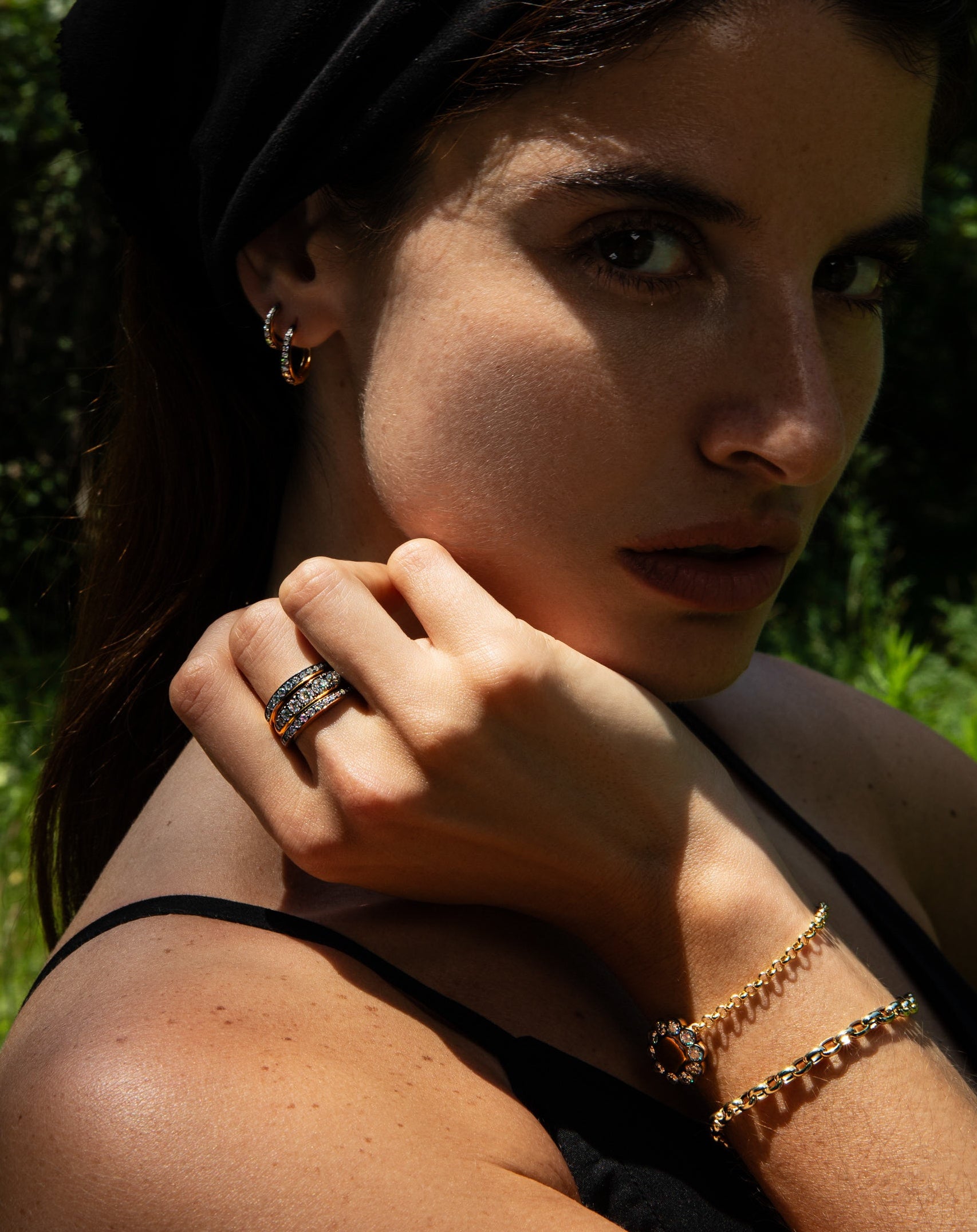 A woman outdoors wearing a black headscarf, black top, and gold jewelry including a ring and bracelets, with a blurred green foliage background.