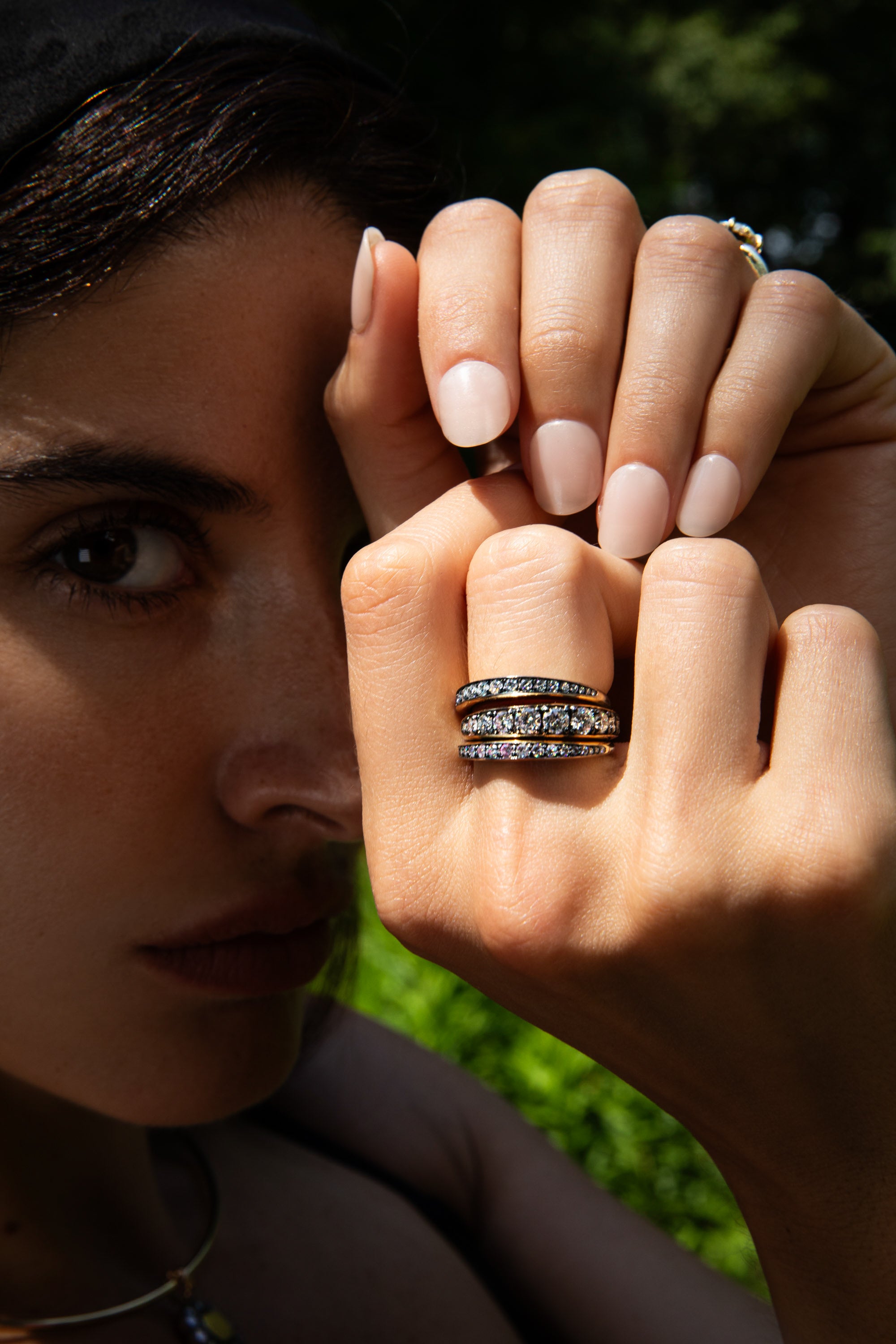 A close-up lifestyle shot of a woman wearing two silver rings set with clear stones, with her hand raised in front of her face against a blurred green outdoor background.