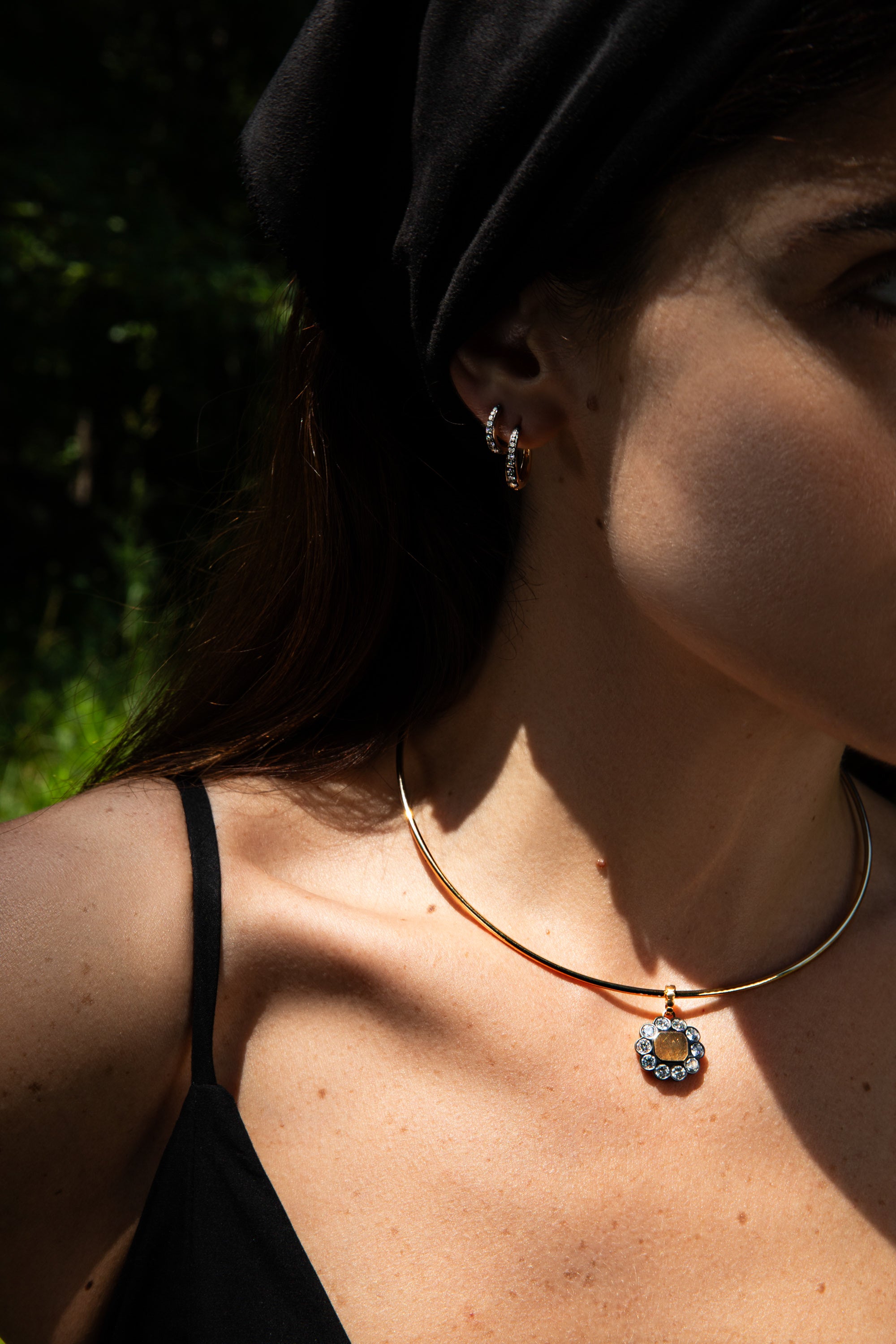 A close-up lifestyle shot of a woman wearing a delicate gold bangle necklace with a silver-toned floral pendant, along with small hoop earrings and a black head covering, set against a dark, blurred outdoor background.