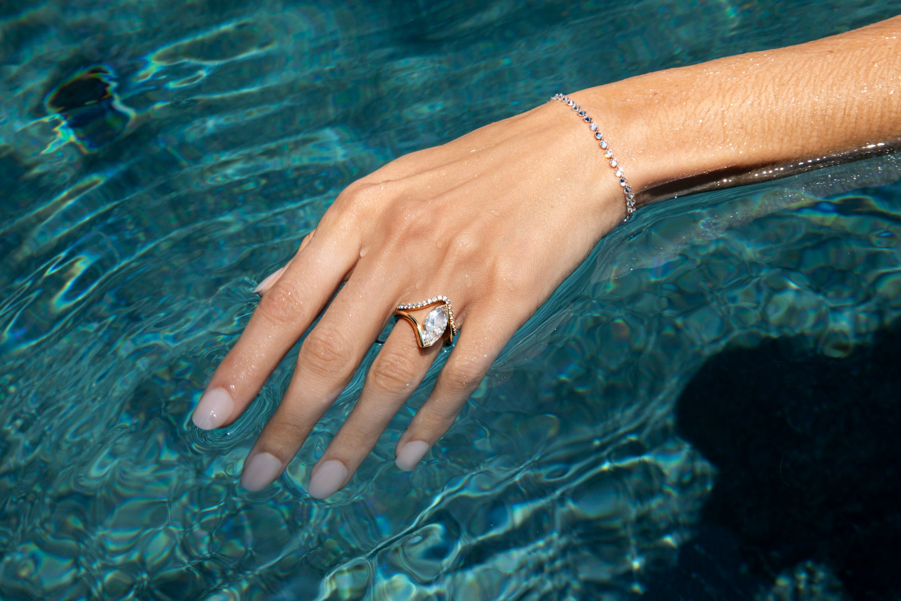 A  close-up of a models hand resting in clear blue water, wearing the cosmic marquise engagement ring, stacked with the cosmic wedding band. Paired with the sparkling pierced diamond tennis bracelet. Shop dripping diamonds for summer.