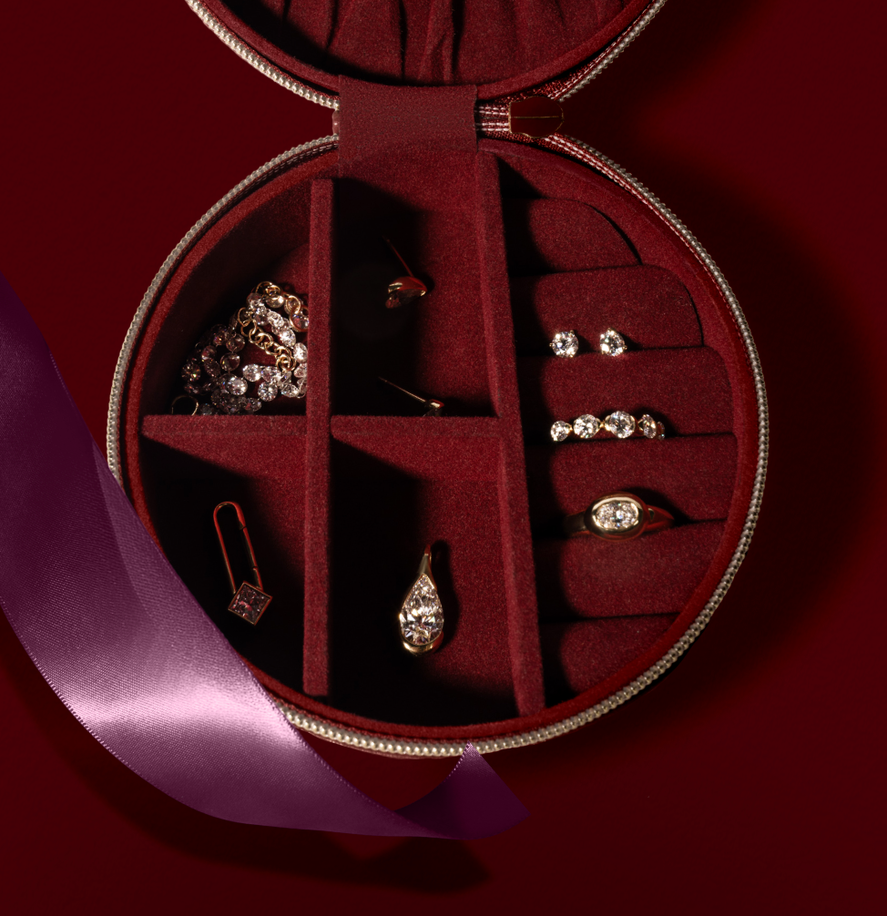 Open round jewelry box with a burgundy velvet interior containing gold and diamond earrings, a ring, a pendant, and a chain necklace, accented by a purple ribbon.