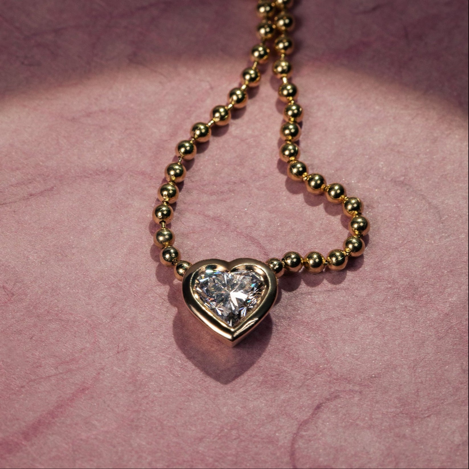 Shop the valentine's day shop and select ready to ship pieces for delivery by 02/14. Gold necklace with a heart-shaped diamond pendant on a pink textured background.