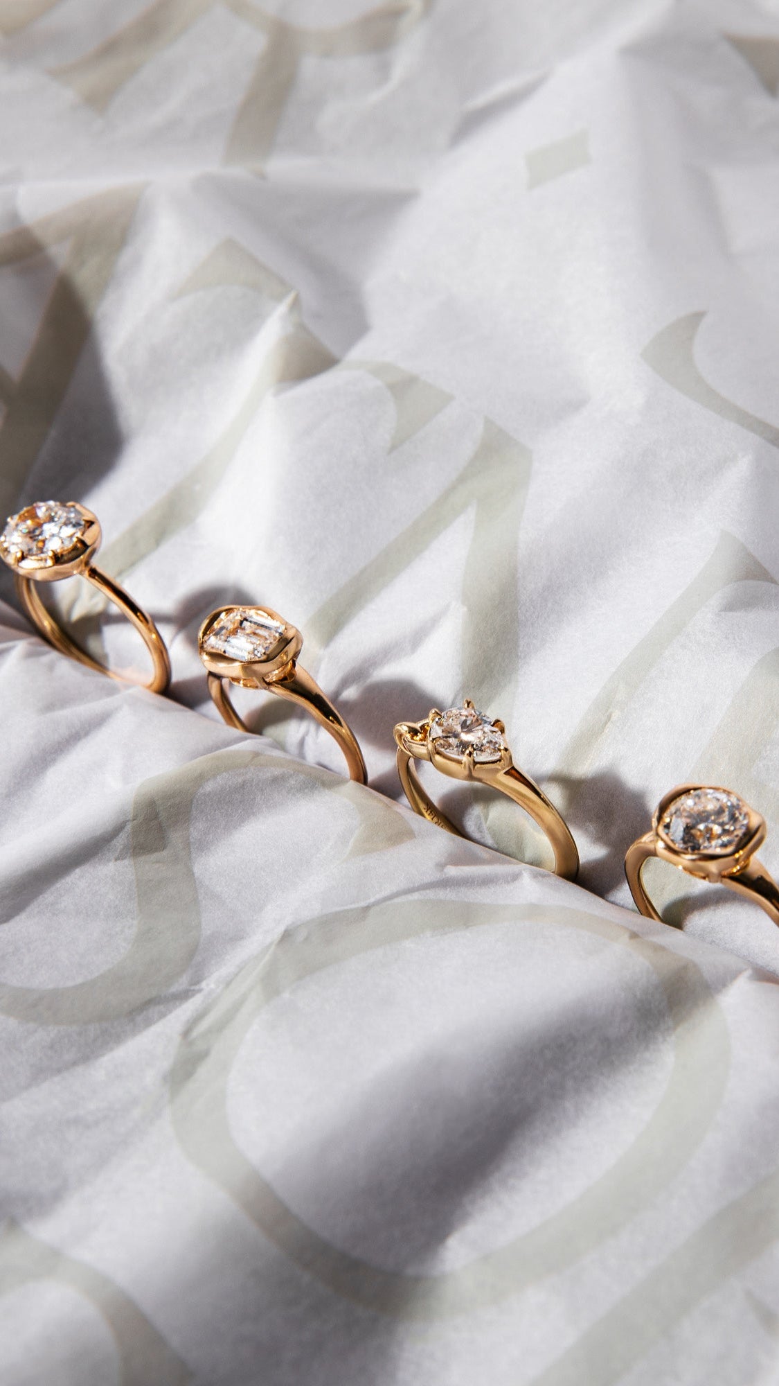 Five yellow gold pinky rings with lab grown diamonds are lined up diagonally on a sophie ratner logo tissue paper.