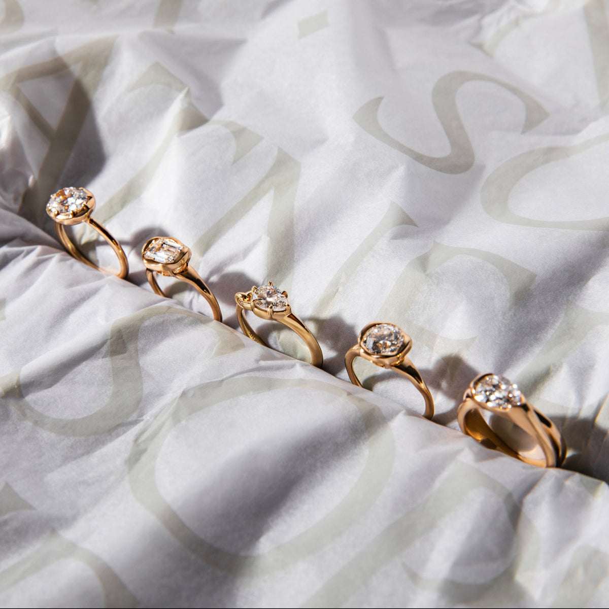 Five yellow gold pinky rings with various lab grown diamond shapes are lined up diagonally on sophie ratner branded tissue paper. introducing new sophie ratner pinky rings. 